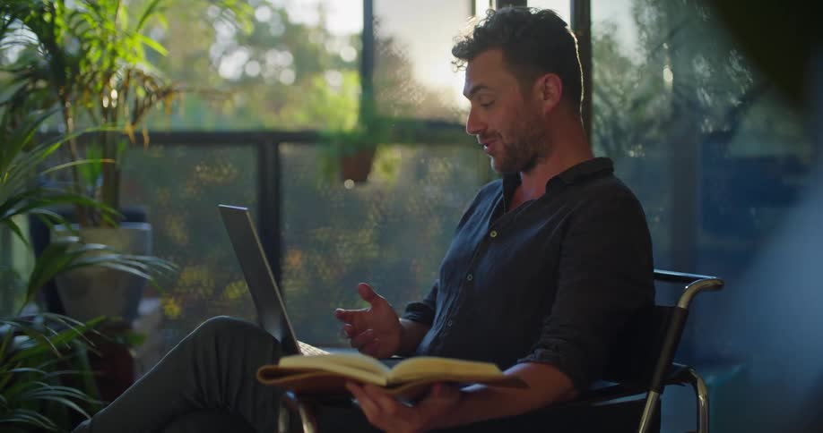A man is sitting in a chair holding a book in one hand and typing on a laptop computer with the other hand