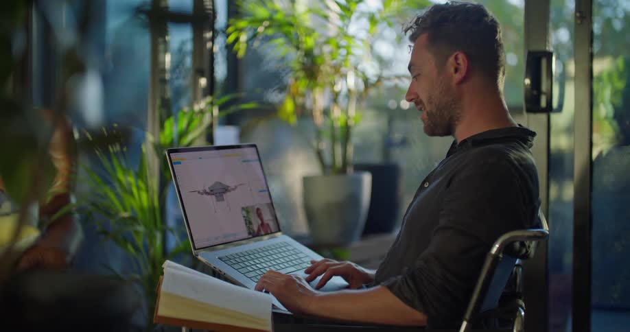 Mna with blue shirt and beard makes video call and works on laptop in living room with many plants