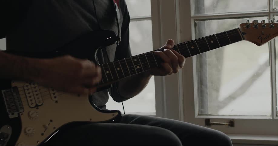 Guitarist sits near window playing electric guitar