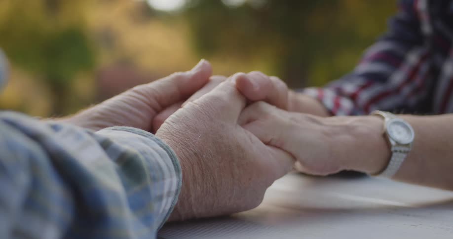 Elderly man and woman wearing tartan shirts sit at table holding hands outdoors