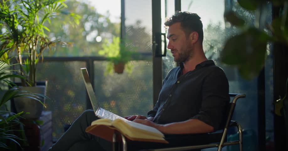 A man is sitting in a chair talking to someone on a laptop computer and his hand is on an open book