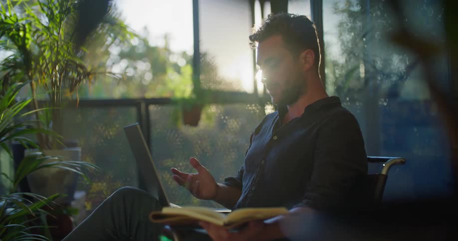 A man sits using his laptop and talking with someone while reading from a book