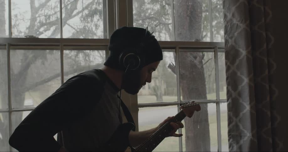A man is near a window wearing headphones and playing a guitar