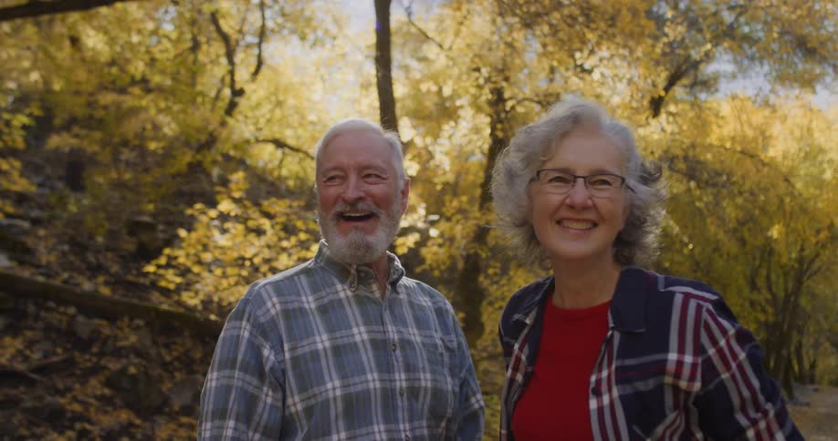 An elderly couple are outside standing together smiling and laughing and there is a forest of trees in bright fall colors behind them