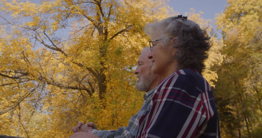 An elderly man and woman hold hands on a bridge in forest with trees covered in fall leaves