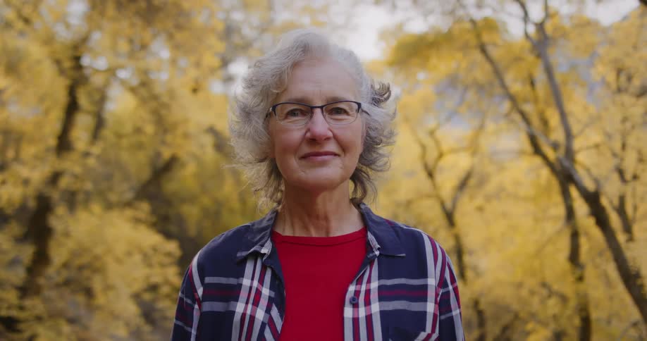 Elderly woman with glasses blue and red tartan shirt smiles at camera in autumn forest