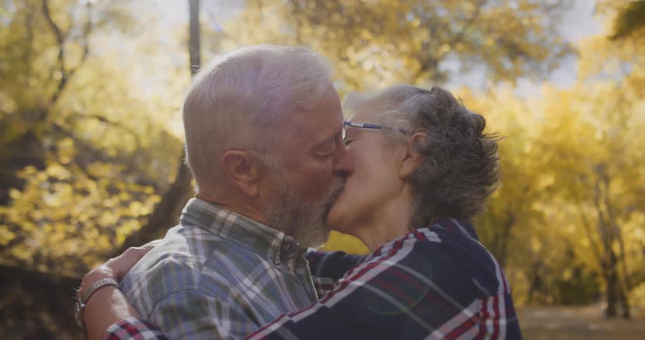 An elderly couple are outside near trees with fall colored leaves and they have their arms around each other and are kissing
