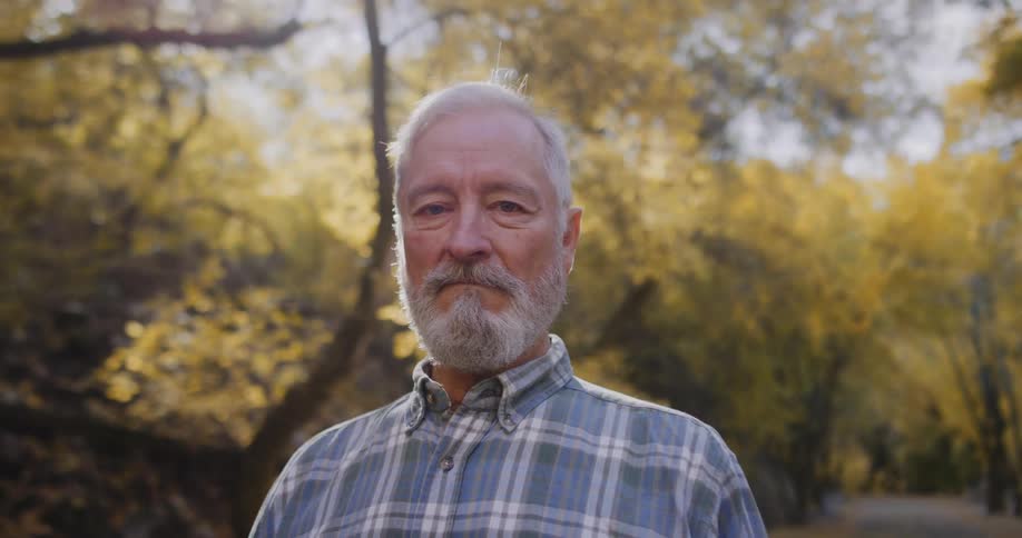Elderly man with beard and tartan shirt looks down at camera in the forest