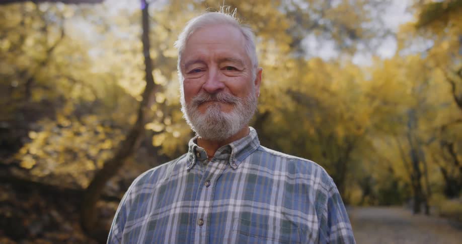 Elderly man with beard and blue tartan shirt smiles at camera in autumn forest
