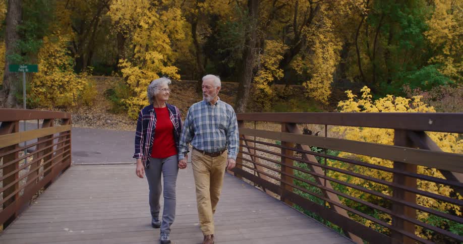 An elderly couple are outside holding hands as they walk across a footbridge and there is a forest of trees in bright fall colors behind them