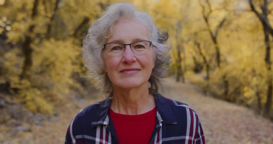 An elderly woman is standing on a dirt road and there are trees with fall colored leaves behind her