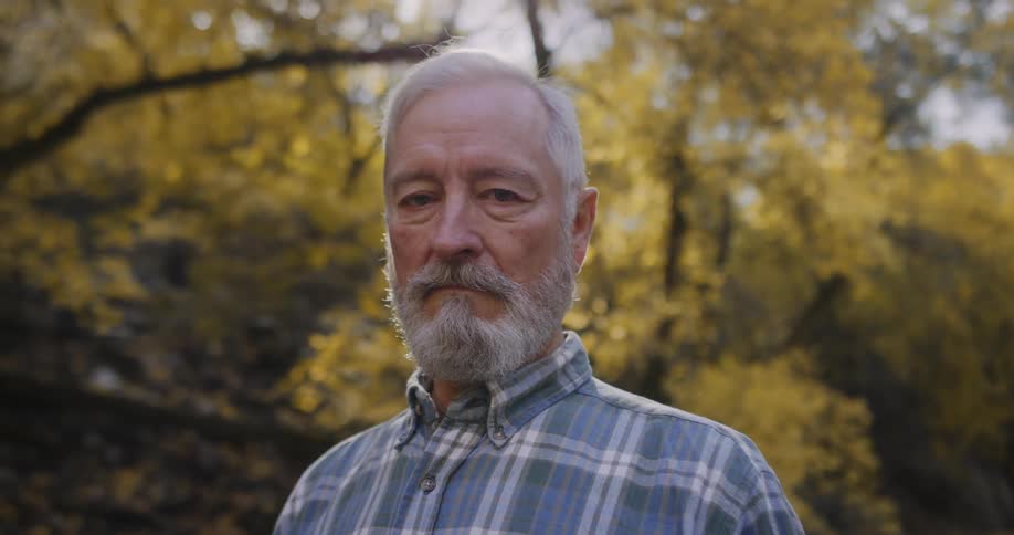 Serious looking man with beard and blue tartan shirt looks at camera in the autumn forest
