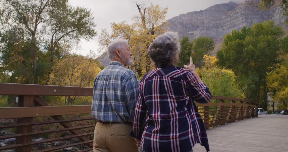 An elderly man and woman walk across a bridge over a river in a forest area with fall leaves