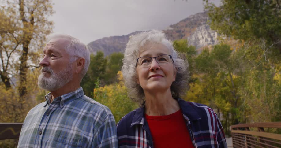 An elderly man and woman walk across a bridge in a forest and look at the fall colors
