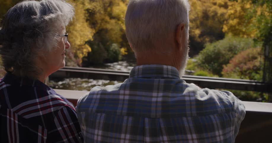 Elderly man with beard and blue tartan shirt and elderly woman with glasses look down from the forest bridge