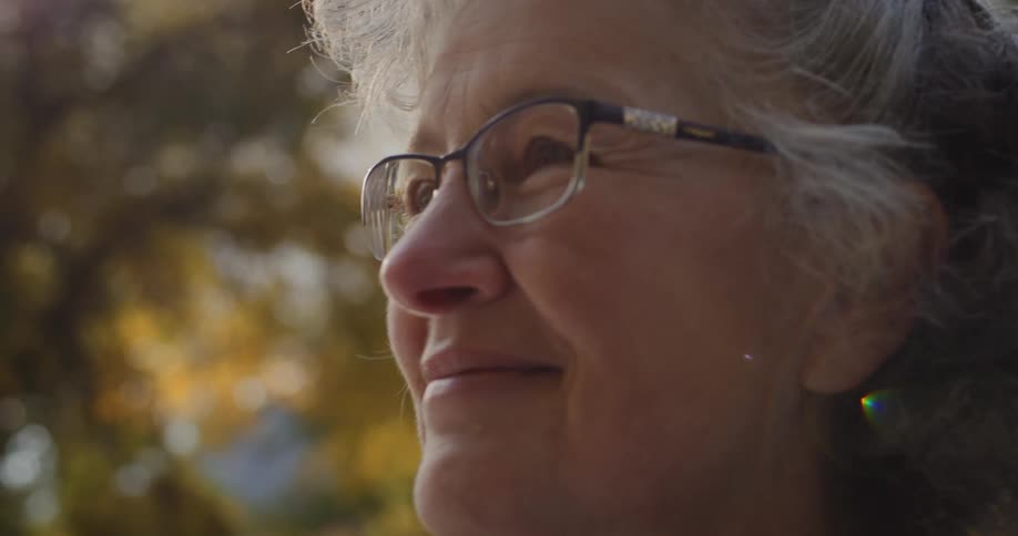 The sun shines on the face of an elderly woman wearing glasses as she stands outside smiling