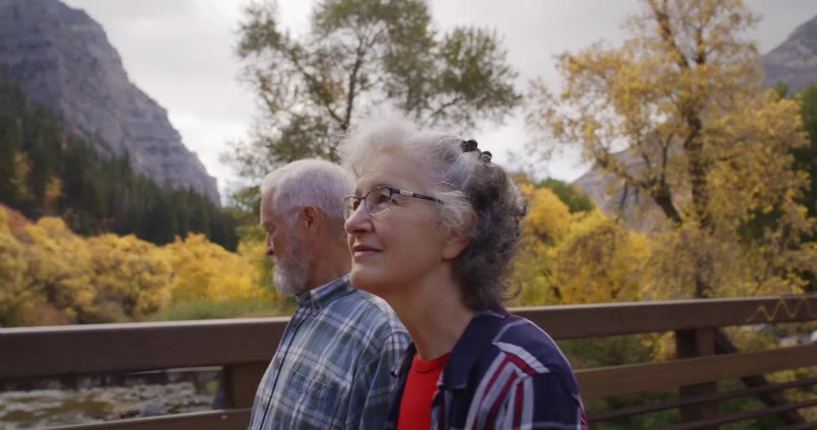Elderly couple hold hands while walking on a bridge over a river near the mountains