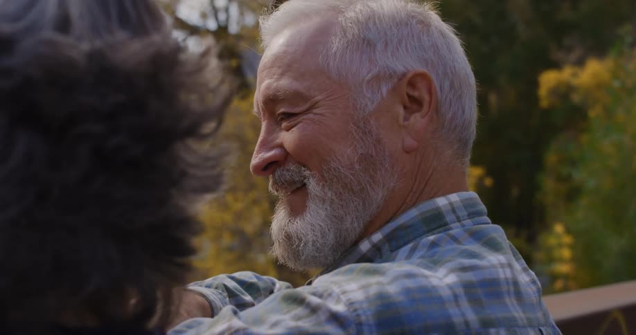 An elderly man smiles and laughs as he talks with a woman in a forest filled with fall trees