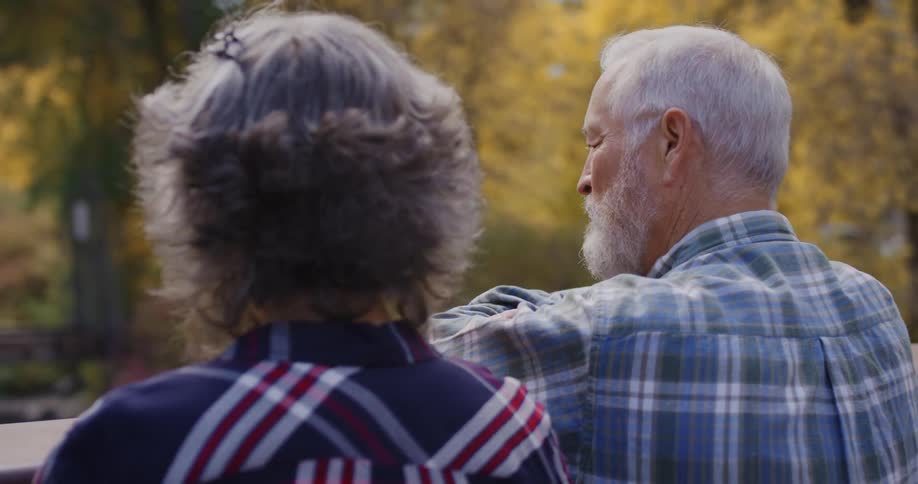 An elderly man and woman stand on a bridge in a forest filled with fall trees and talk