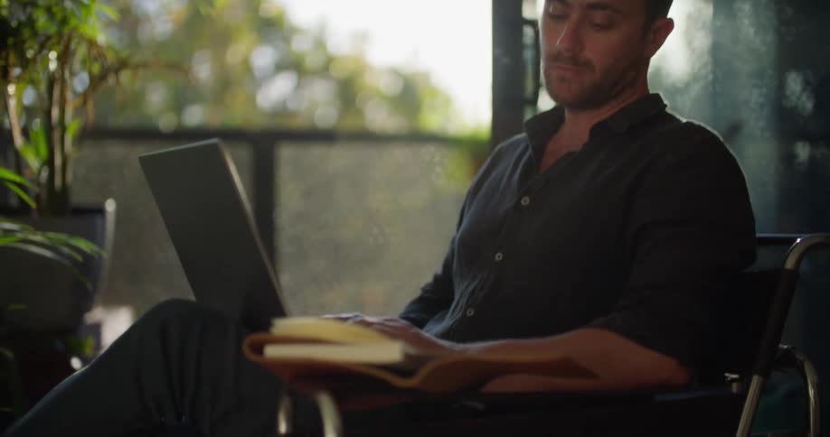 A man is sitting in front of an open balcony door typing on his computer