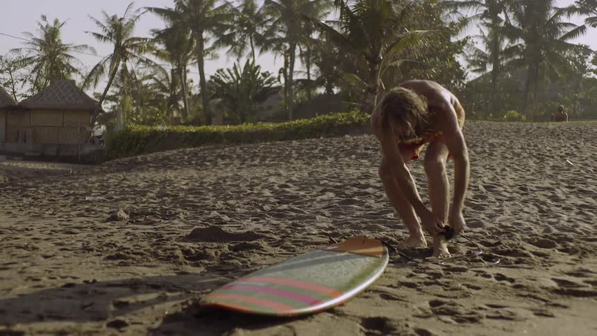 A bearded man ties a surfboard to his ankle