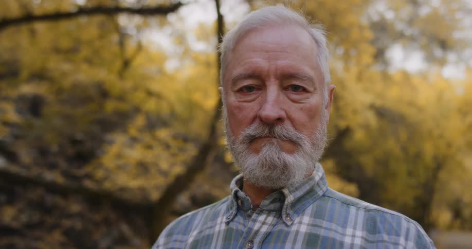 Close up of a bearded mature man with white hair looking at the camera in the forest
