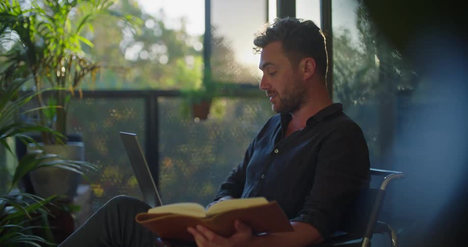 A young man sits at home with a laptop on his knee and a book in his hand as he video chats on the compute while looking at the book