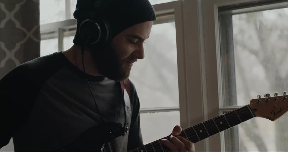 A man is near a window wearing headphones and playing a guitar