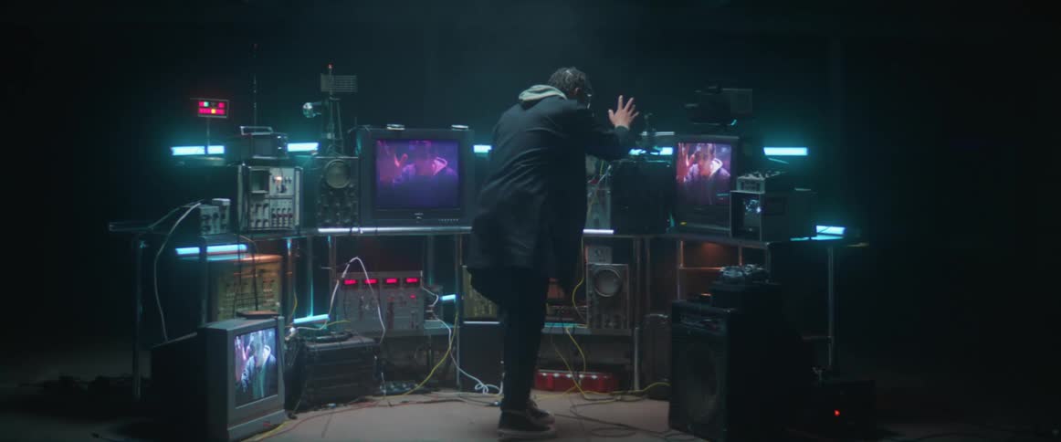 A man walks into a dark room filled with televisions and electronics and he sees himself on the screens