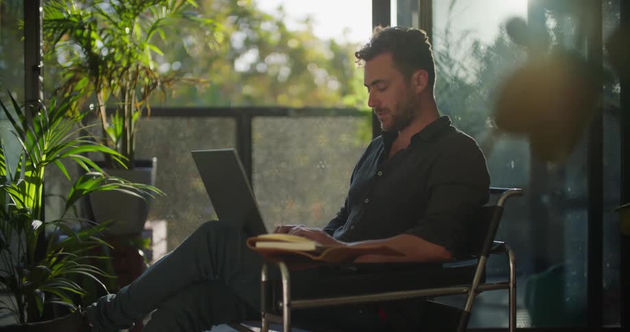 Man sitting on a chair in the living room reads on a notebook and writes on a laptop at morning