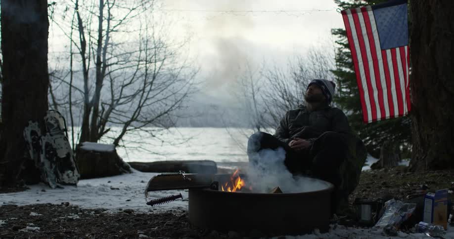 A man sits near a campfire with an american flag behind him