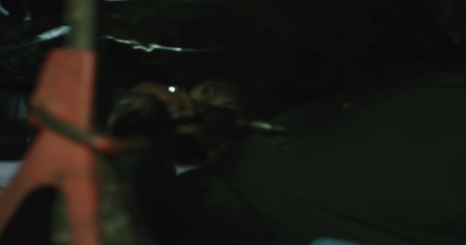 A man wearing a headlamp picks up a drill and moves underneath a car