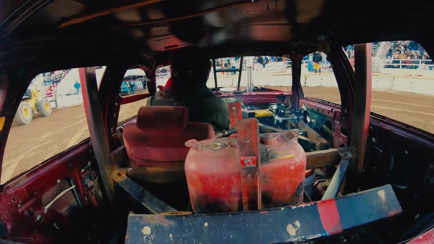 Person drives car around dirt arena as camera man stands with tripod near audience