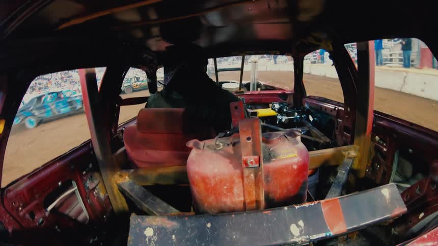 Demolition derby driver maneuvers car around dirt arena as crowd watches