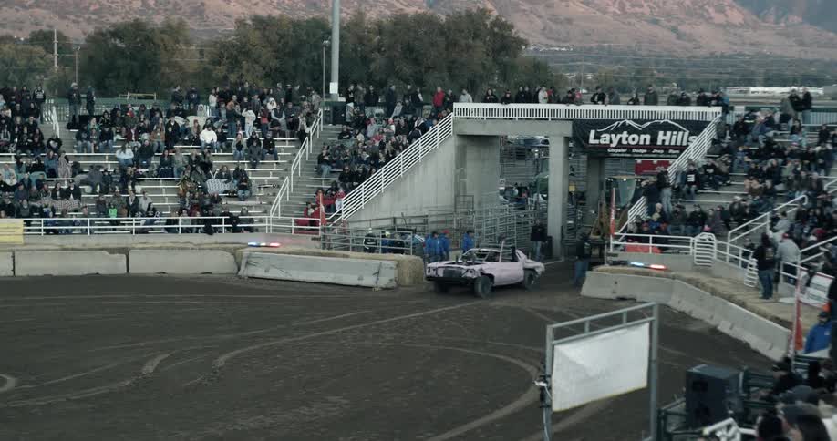 Lights flash as a car enters a demolition derby stadium with people in the stands