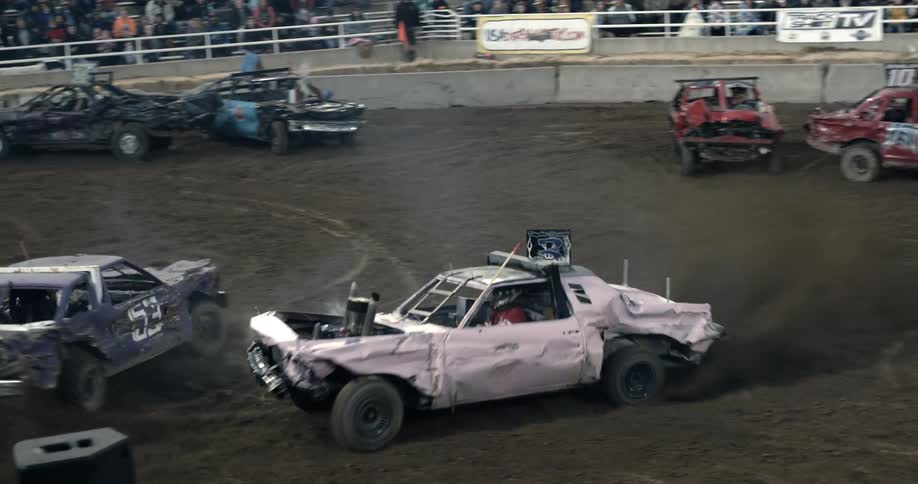 Cars drive along demo derby arena crashing into each other as the audience watches