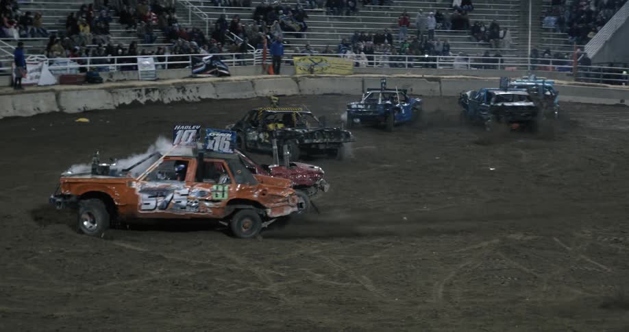 Audience watches from the stands at night as demo derby contestants drive around dirt arena crashing into each other