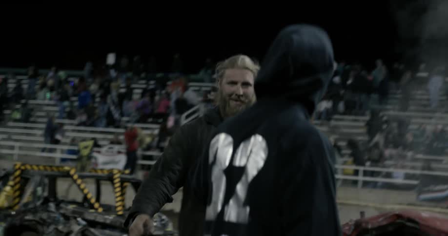 A man jumps out of a wrecked car in a demolition derby as a crowd in the bleachers watch