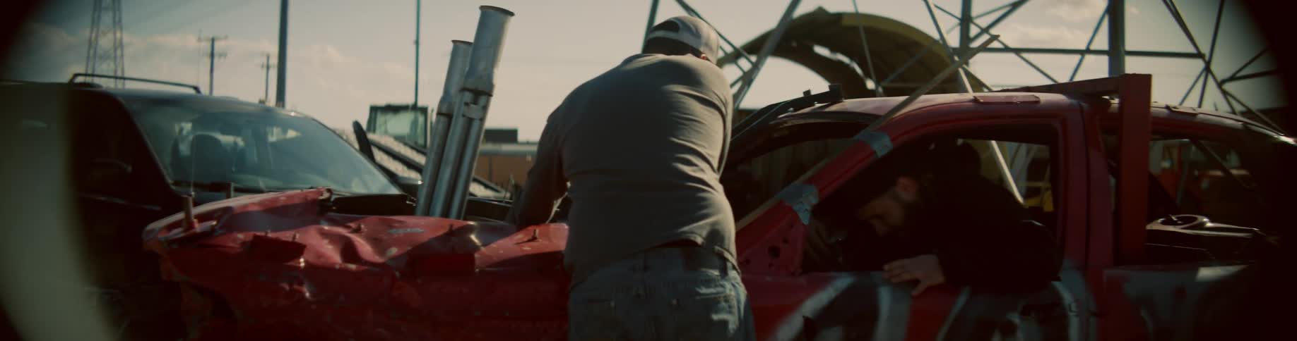 A man climbs into a car through the driver side window as a friend finishes adjusting the engine for a demolition derby