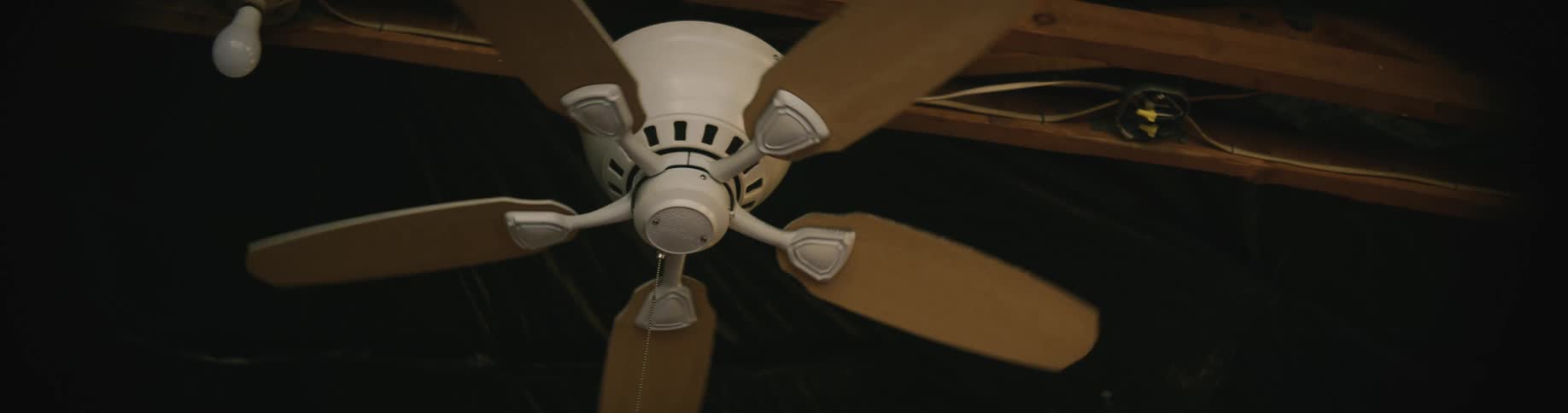 A ceiling fan rotates slowly next to a naked bulb with wires running between beams