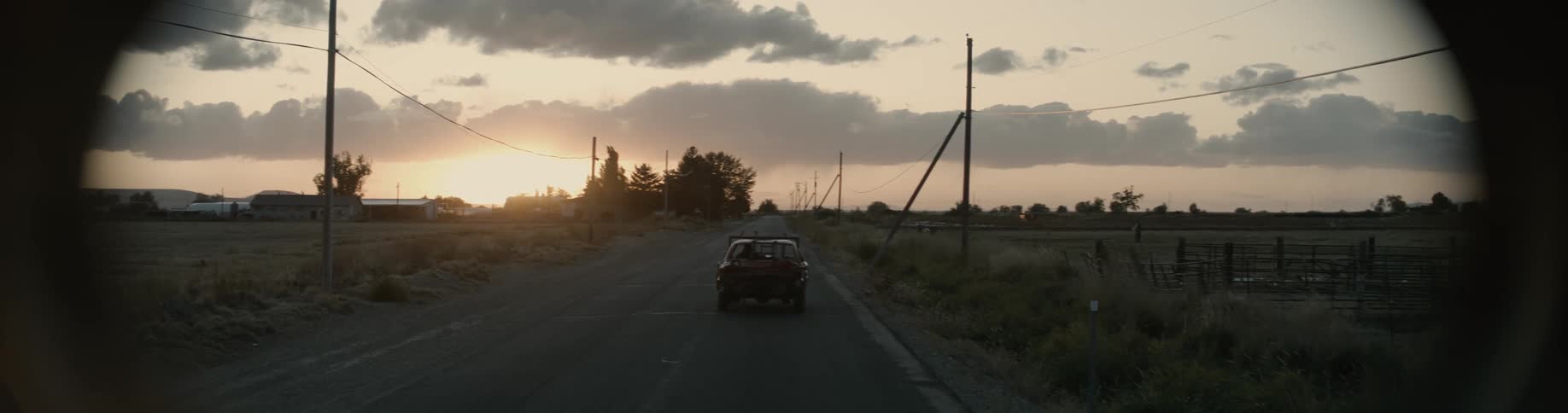 An automobile travels on a rural road as the sun sets on the horizon in a cloudy filled sky