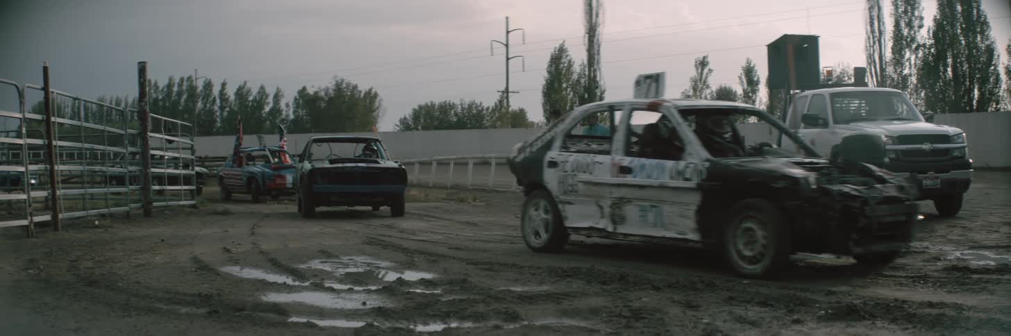 Two men are standing near a fence waving as cars are moving into the demolition derby track