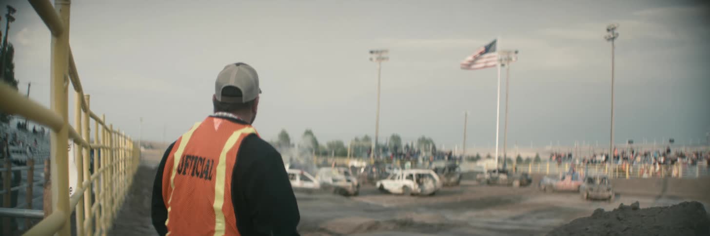 Demo derby official stands watching competing vehicles maneuver in dirt as crowd fills stands