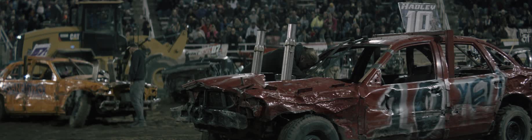 Men are inspecting a smashed car in a demolition derby as the people in the stands watch what is happening