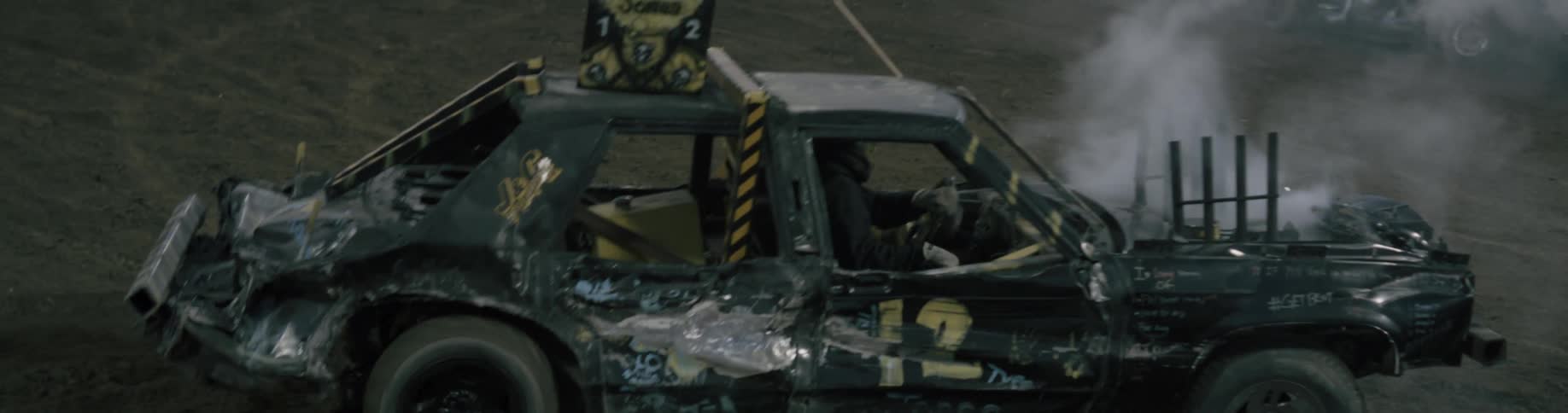 An audience in the stands watches as cars crash into each other on a dirt track in a demolition derby