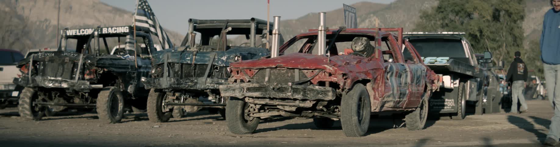 Men walk around past cars parked and ready for a demolition derby