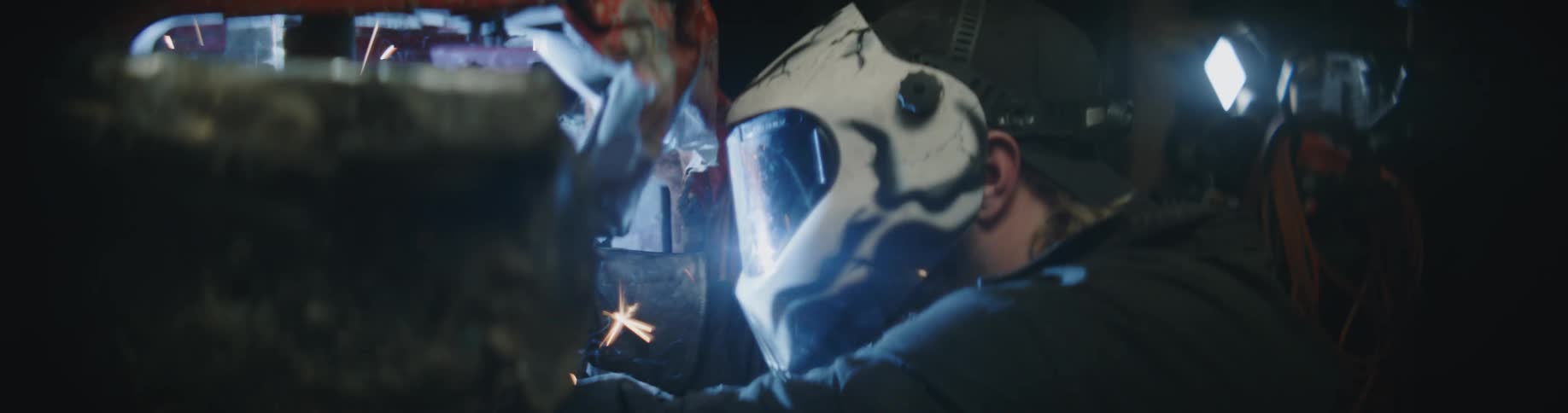 A man is wearing a welding helmet and light shines as sparks fly from the car he is welding