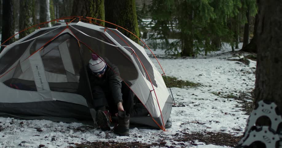 A man unzips his tent and puts his boots on before exiting