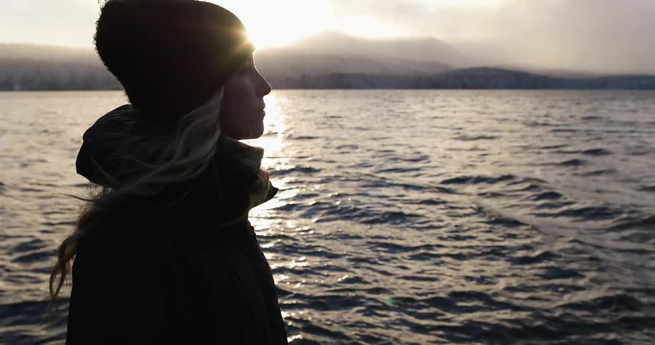 A woman is standing by the water on a windy day at sunset