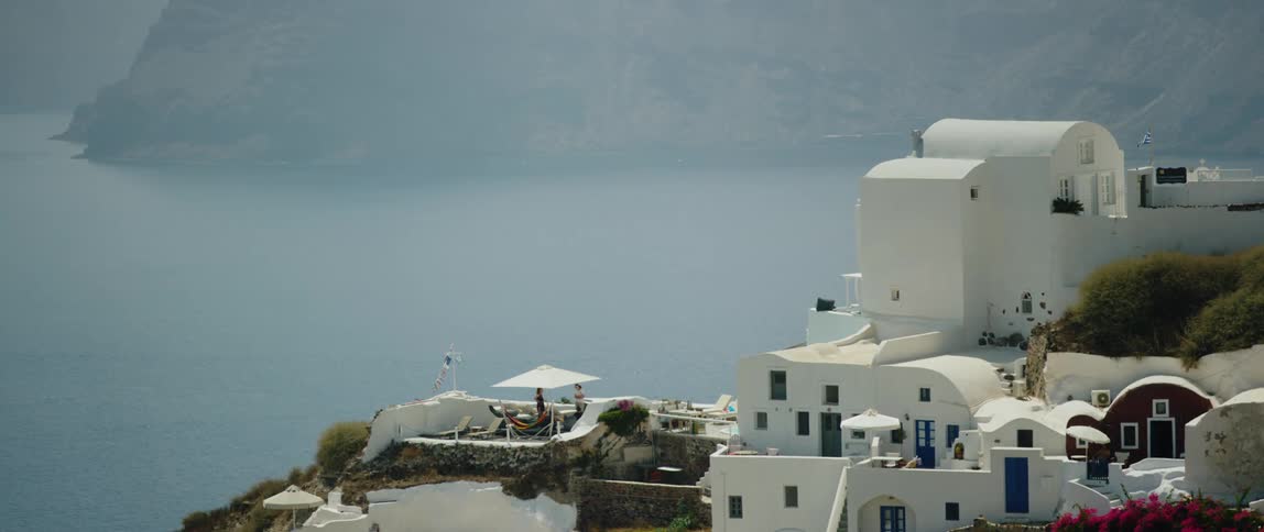 White buildings sit on a cliff overlooking a large body of water and people are talking on a balcony